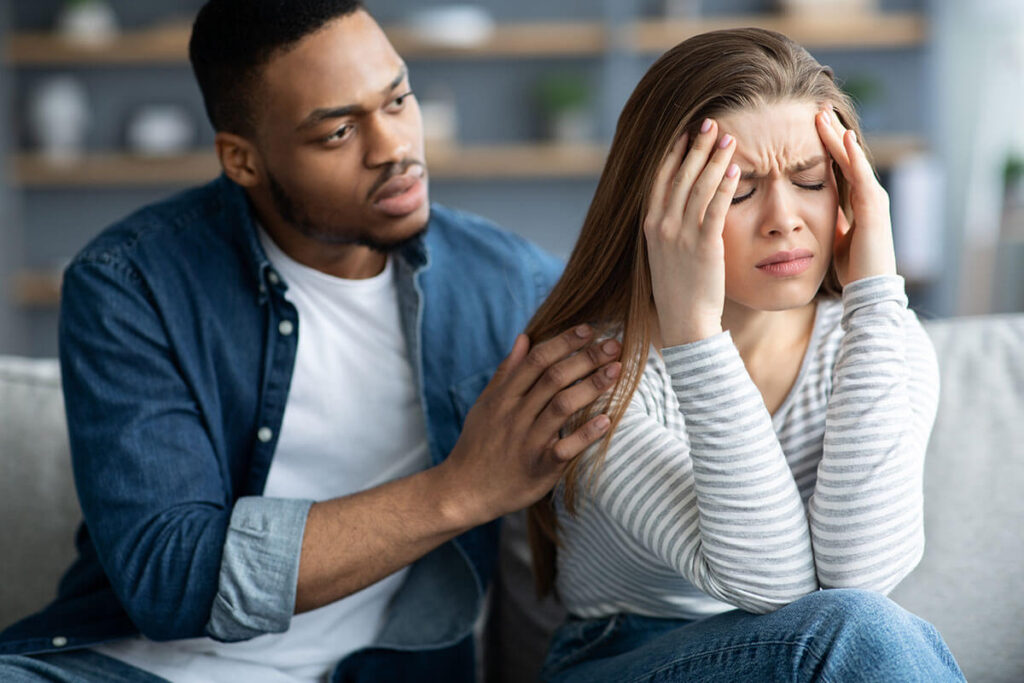 A man sits beside a woman who looks stressed, holding her head with her hands while he places his hands on her shoulders in a comforting gesture.