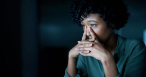 A person with curly hair rests their face against their hands in a dimly lit environment, appearing contemplative or stressed.