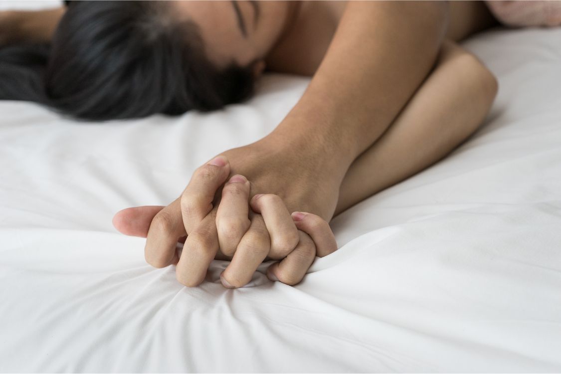 Two people lying on a bed hold hands, their fingers interlocked.