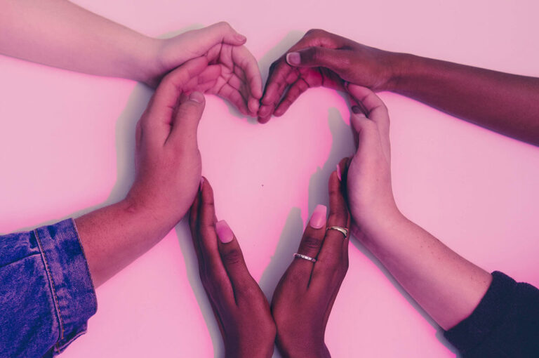 Four pairs of diverse hands form the shape of a heart against a pink background.